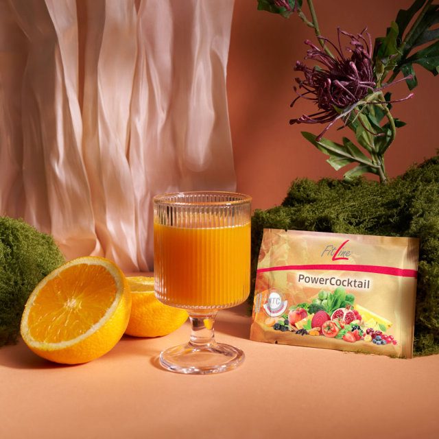 Produktdose PowerCocktail auf hellem Holzuntergrund, warmes Morgenlicht, daneben ein Glas mit gemischtem Getränk in Orange-Ton