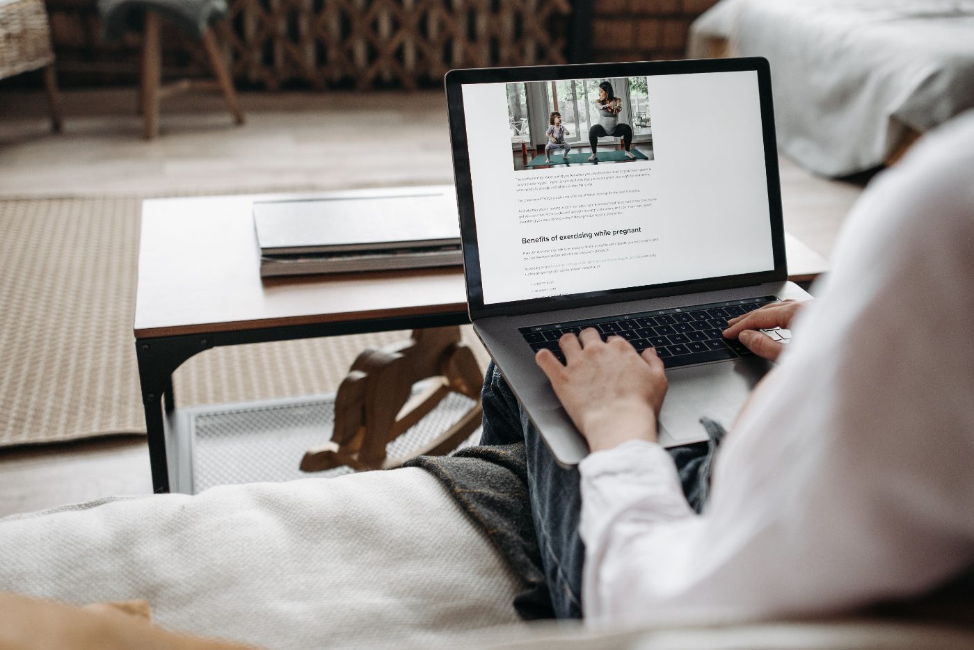 Junge Mutter Mitte 30, sitzt auf dem Sofa mit schlafendem Kind auf dem Arm, Laptop aufgeklappt daneben, ruhiges warmes Licht — der Moment zwischen Pflege und eigenen Plänen